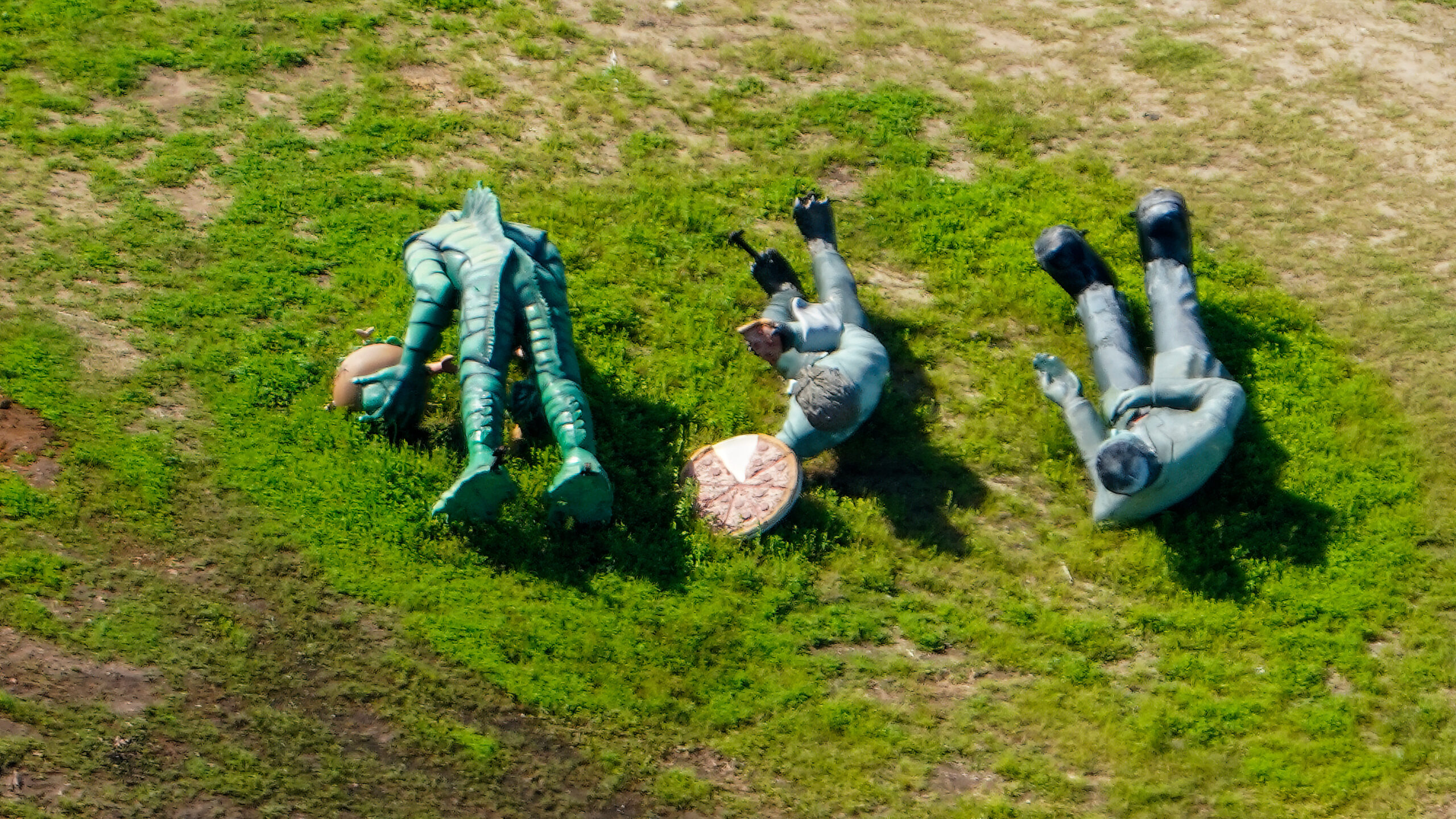 Three life-size creature statues lying in the grass beside a round prop resembling an oversized pizza