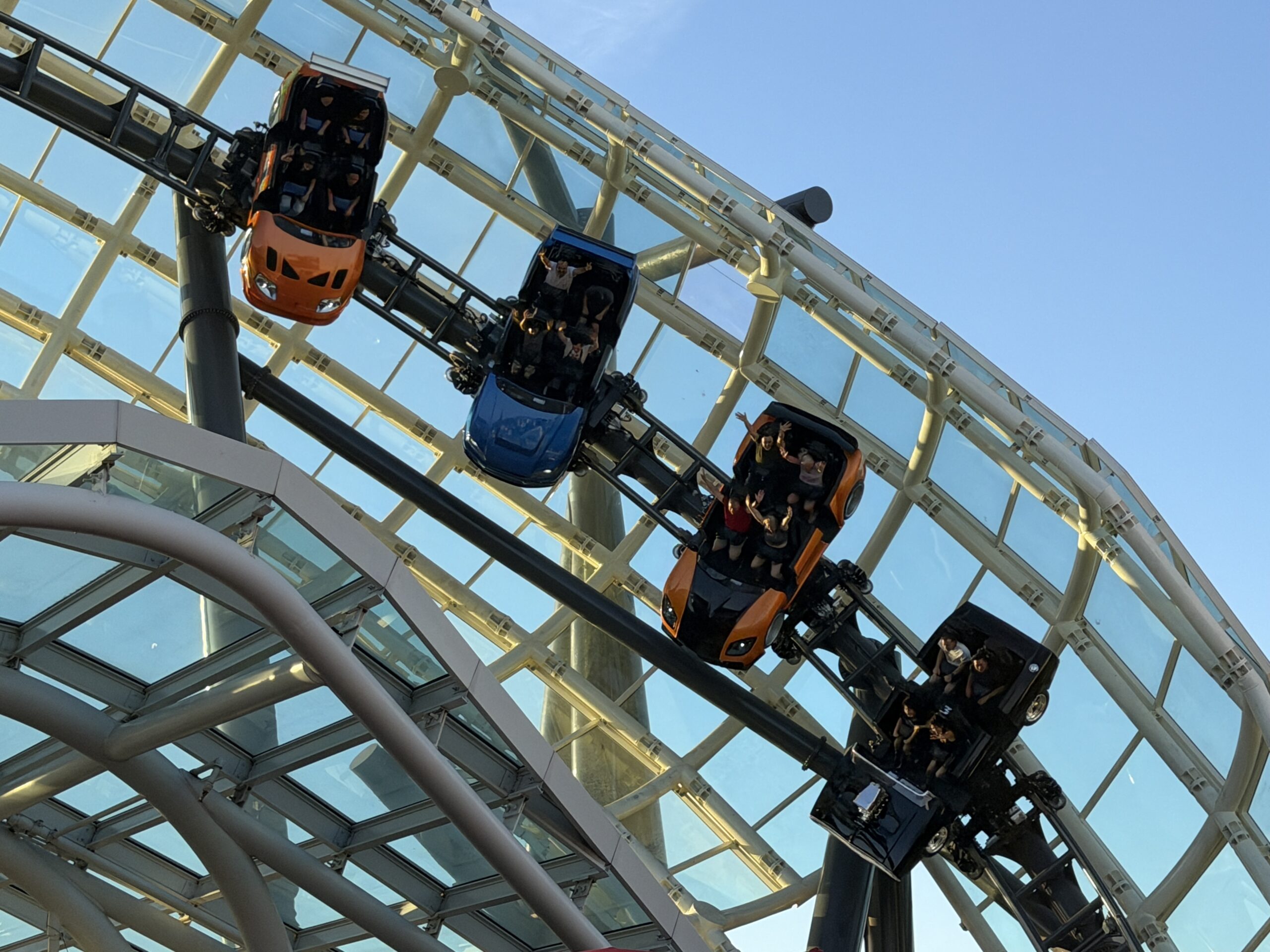 Four car shaped roller coaster vehicles in orange, blue and black mounted on arms beside an elevated track, with riders raising their arms, against a glass and white steel building