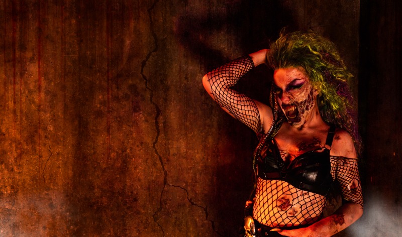 A performer lit in orange against a cracked, stained wall, one arm raised into wild yellow green curls, one cheek and jaw decayed down to exposed teeth