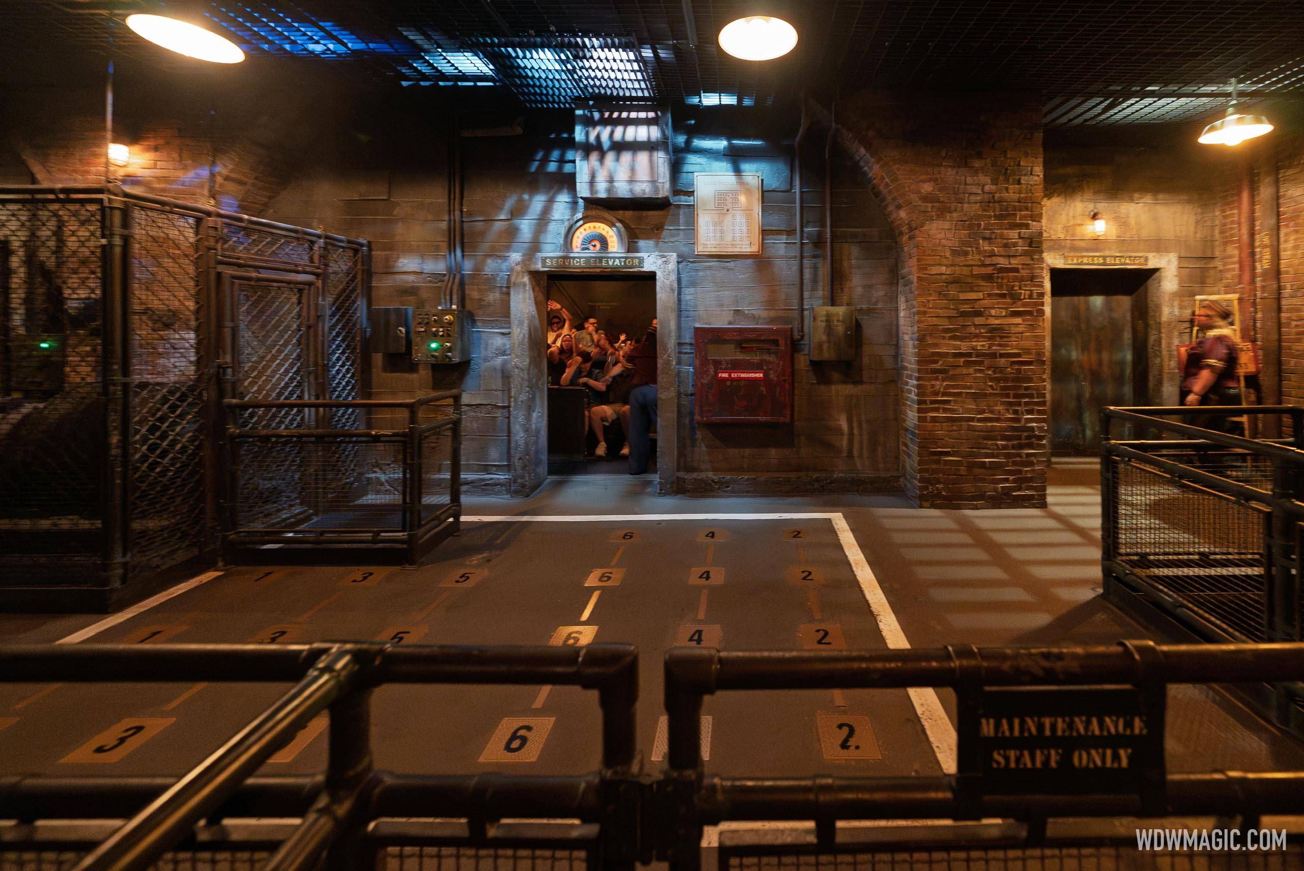Wide view of a drop ride load area from behind a railing, numbered floor row markers leading to an open elevator car with seated guests inside, and a Maintenance Staff Only sign in the foreground
