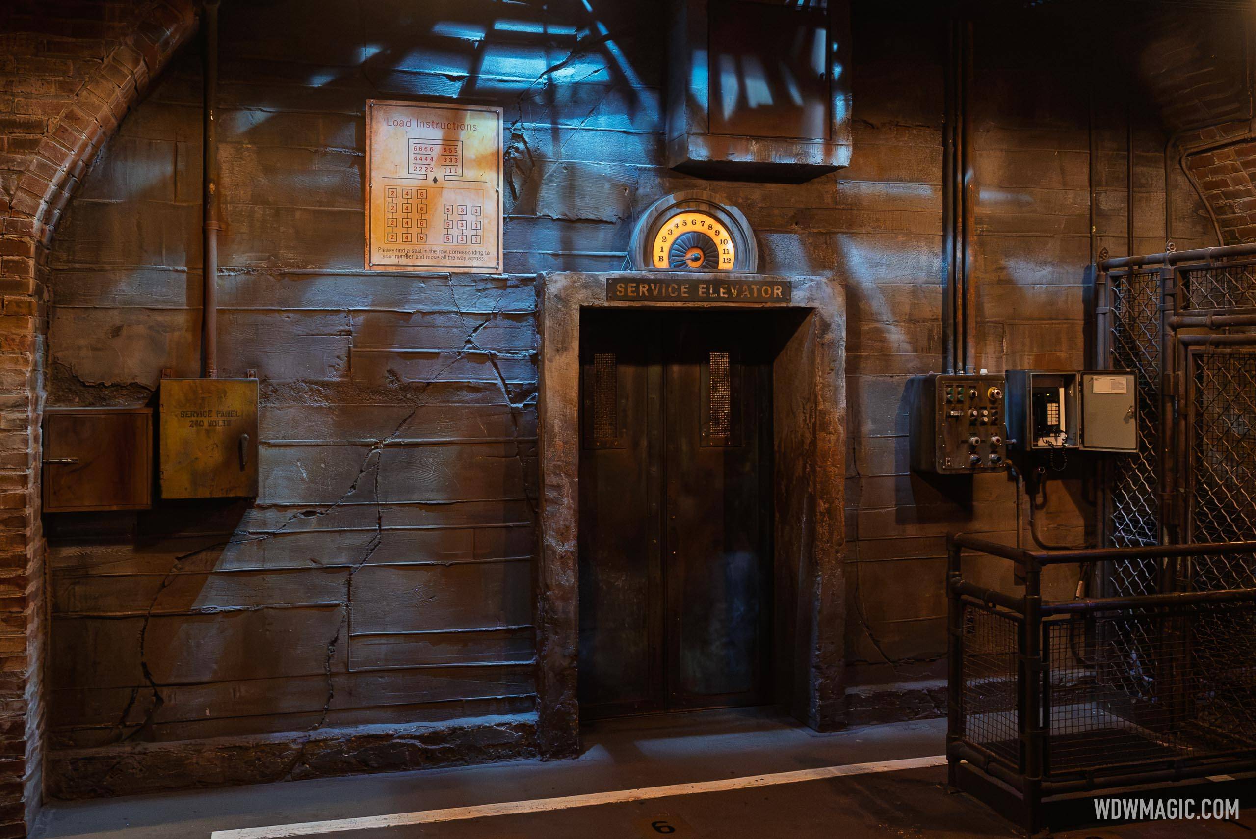 The dimly lit boiler room boarding area of a drop ride, with a lit SERVICE ELEVATOR doorway, a glowing floor indicator dial above it, and a Load Instructions seating placard on the cracked concrete wall