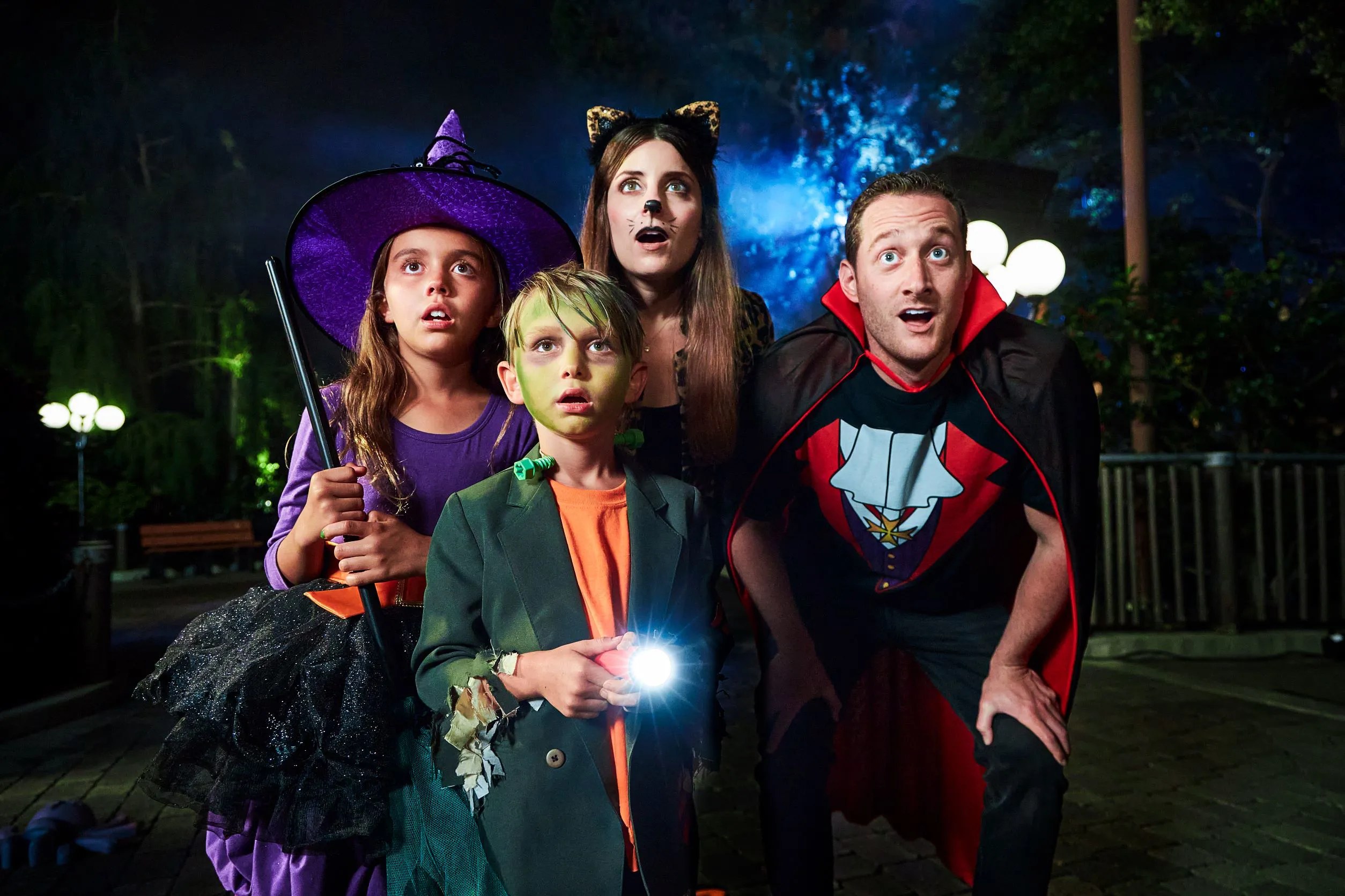 A family of four in Halloween costumes at night in a theme park, a girl in a purple witch hat, a boy with green face paint holding a flashlight, a woman with cat ears and a man in a vampire cape, all looking upward