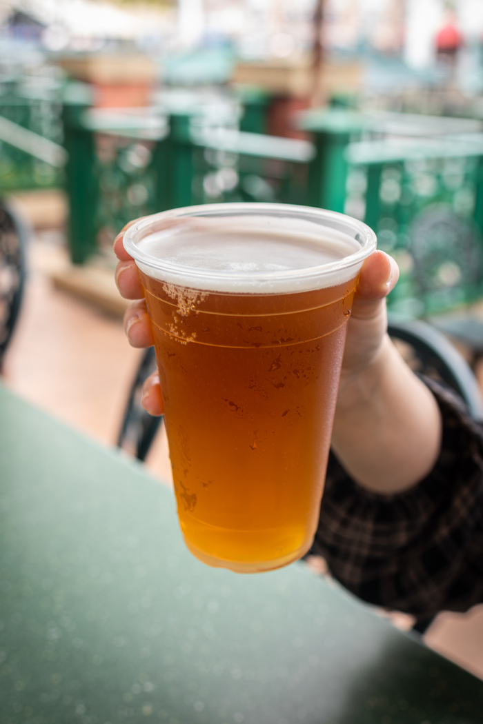A hand holds up a clear plastic cup of amber Dufftoberfest beer with a white head, in front of the green and white Duff Brewery beer garden at Universal Studios Florida