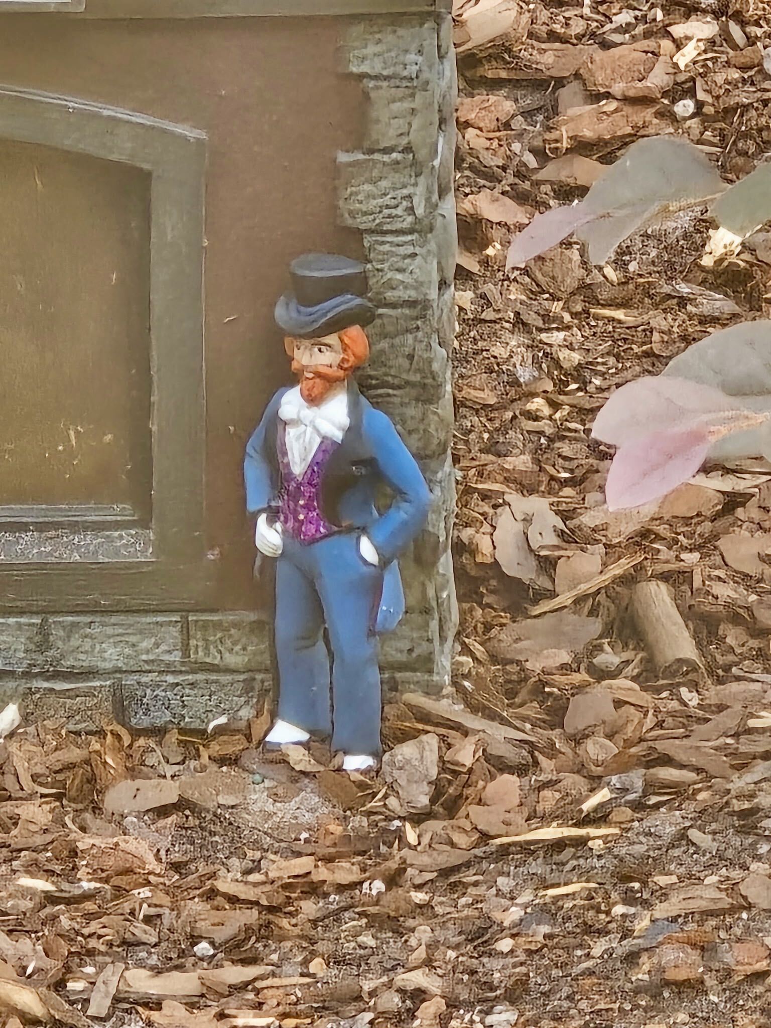 A close up of the miniature Dreamfinder figure in the Germany Train Garden, wearing a blue suit, a grey blue top hat, a magenta patterned vest and a white cravat, with a red orange beard, one hand on his hip, standing on mulch beside a brown building wall with a recessed arched panel and grey stone corner blocks