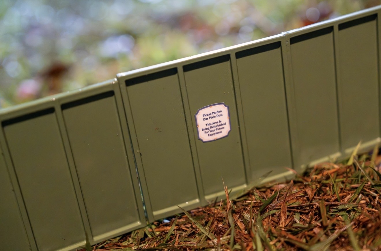A run of miniature olive green construction wall panels in the Germany Train Garden, one of them carrying a small printed sign reading Please Pardon Our Pixie Dust, This Area Is Being Refurbished For Your Future Enjoyment, with mulch and grass along the bottom of the frame