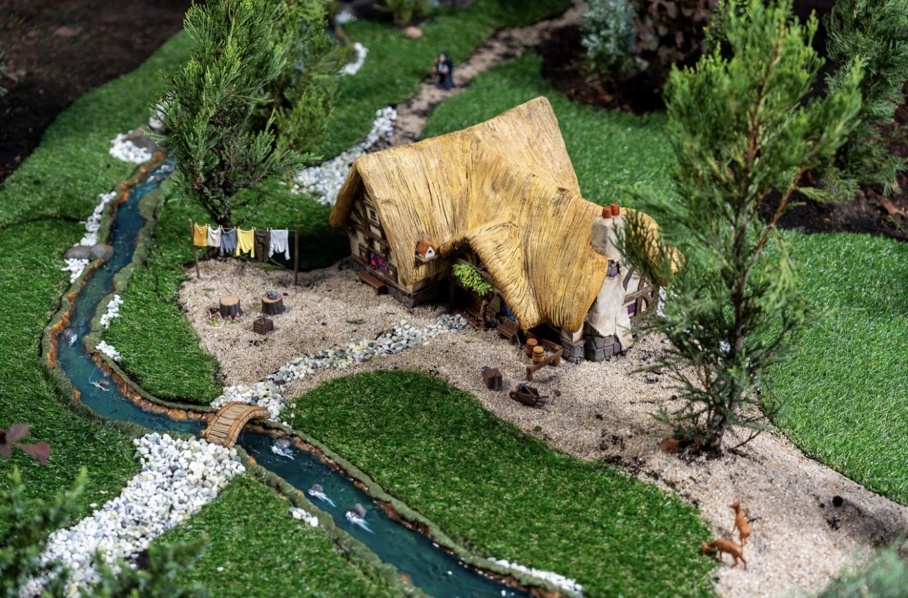 The miniature Snow White cottage in the Germany Train Garden, with a yellow thatched roof, a clothesline strung with tiny laundry, tree stump stools, a teal creek crossed by a small wooden footbridge, two orange deer at lower right and a small dark cloaked figure on the path at the top