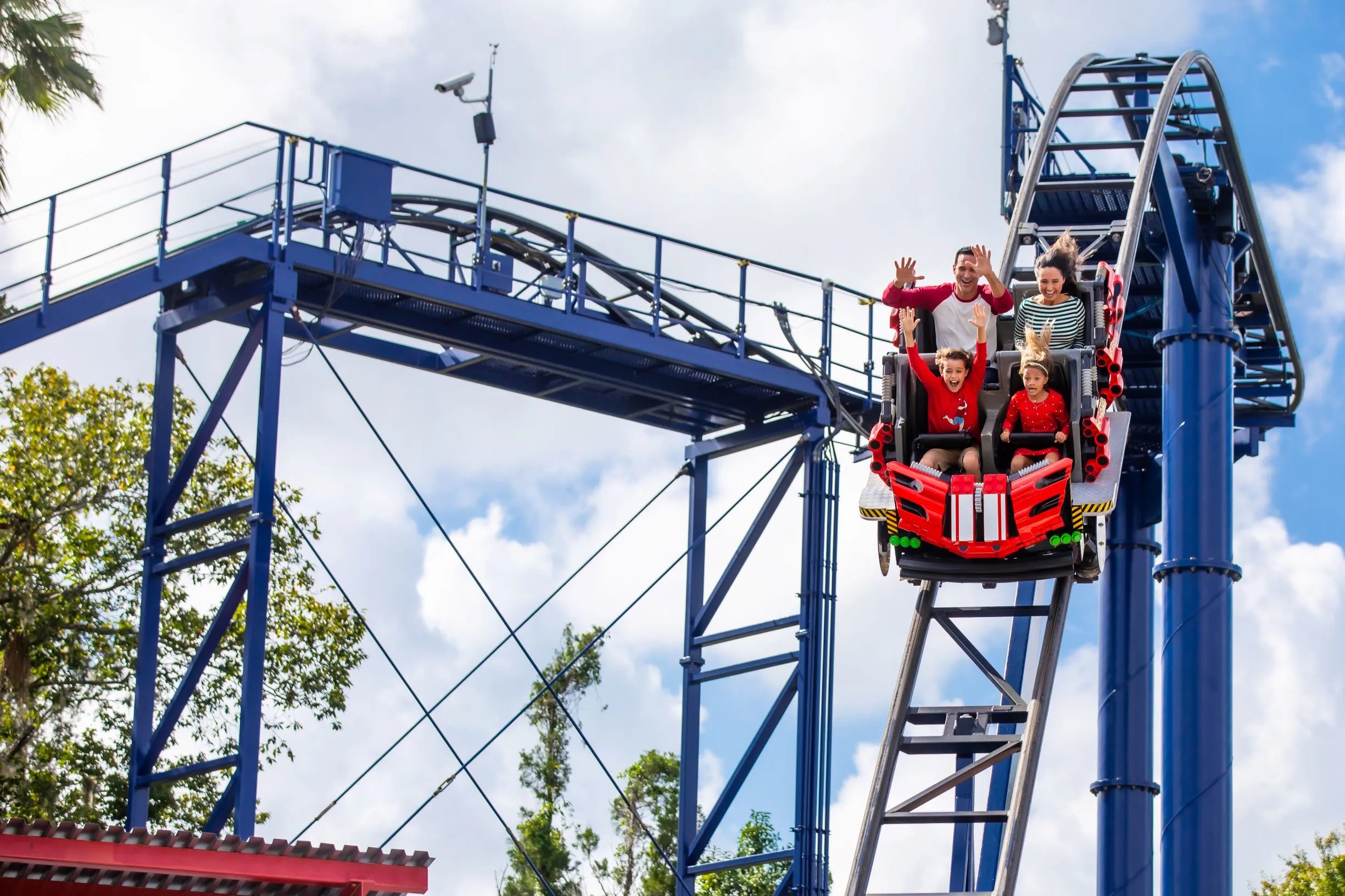 A family of four riding a red LEGO themed coaster car, the two adults with hands raised, on grey steel track through the ride's blue steel structure, against a partly cloudy sky