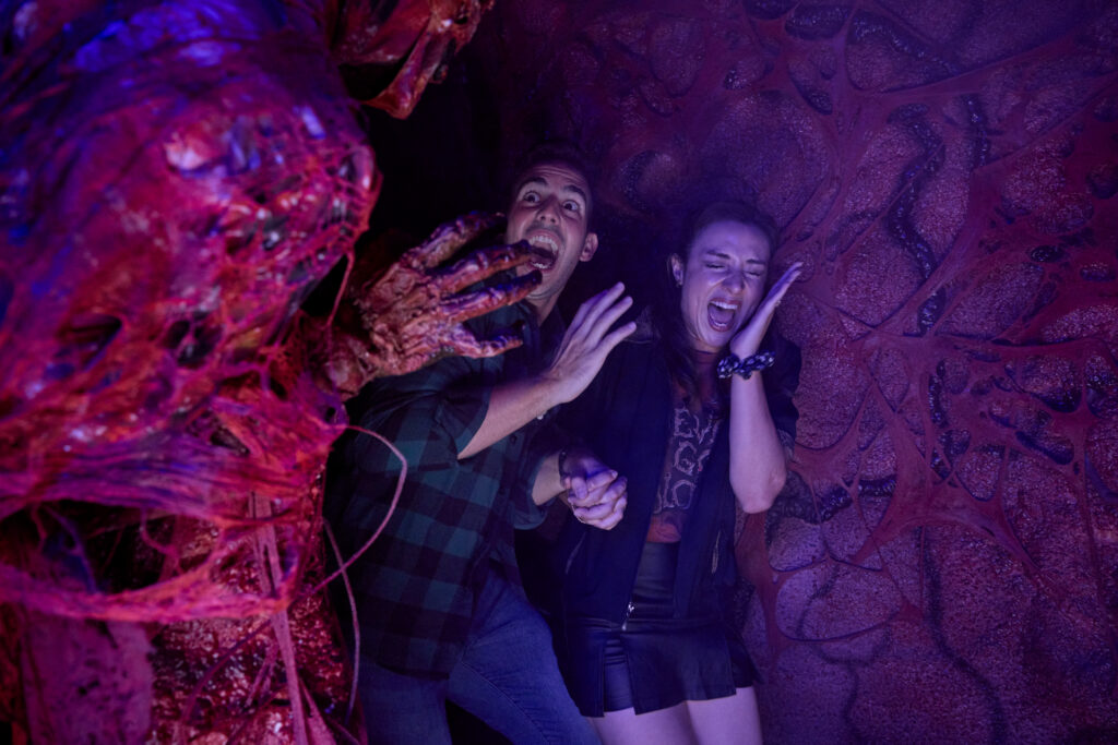 Generic Halloween Horror Nights publicity photo, not from any of the three houses in this story: two guests scream in a haunted house corridor with fleshy red walls, lit purple and magenta, as a clawed hand reaches toward them.