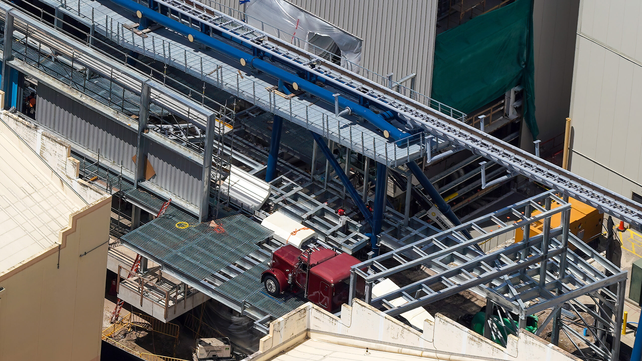 An aerial view of the exposed silver steel framework of the Hollywood Drift first scene, with blue coaster track on supports crossing the upper portion of the frame, the burgundy truck cab on grating below and a green construction screen at right