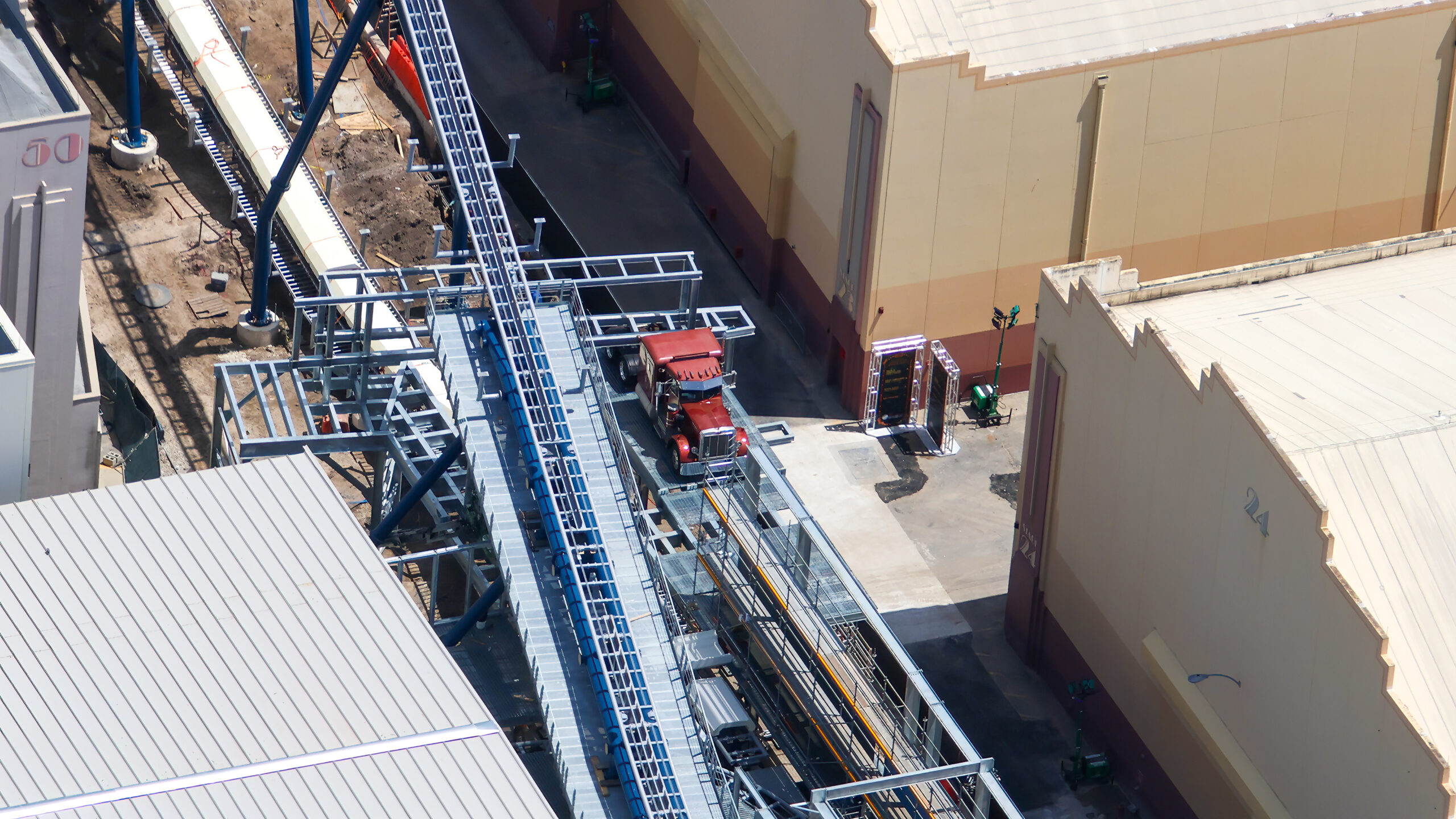 A wide aerial view of the Fast & Furious: Hollywood Drift construction site at Universal Studios Florida, with blue coaster track running diagonally next to a silver steel catwalk, the burgundy truck cab on a grated platform alongside, and tan soundstage walls to the right