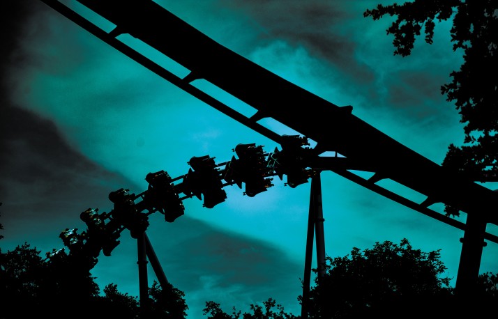 A roller coaster train silhouetted in black against a teal green night sky, tilted hard on a curving section of track, framed by dark tree canopy