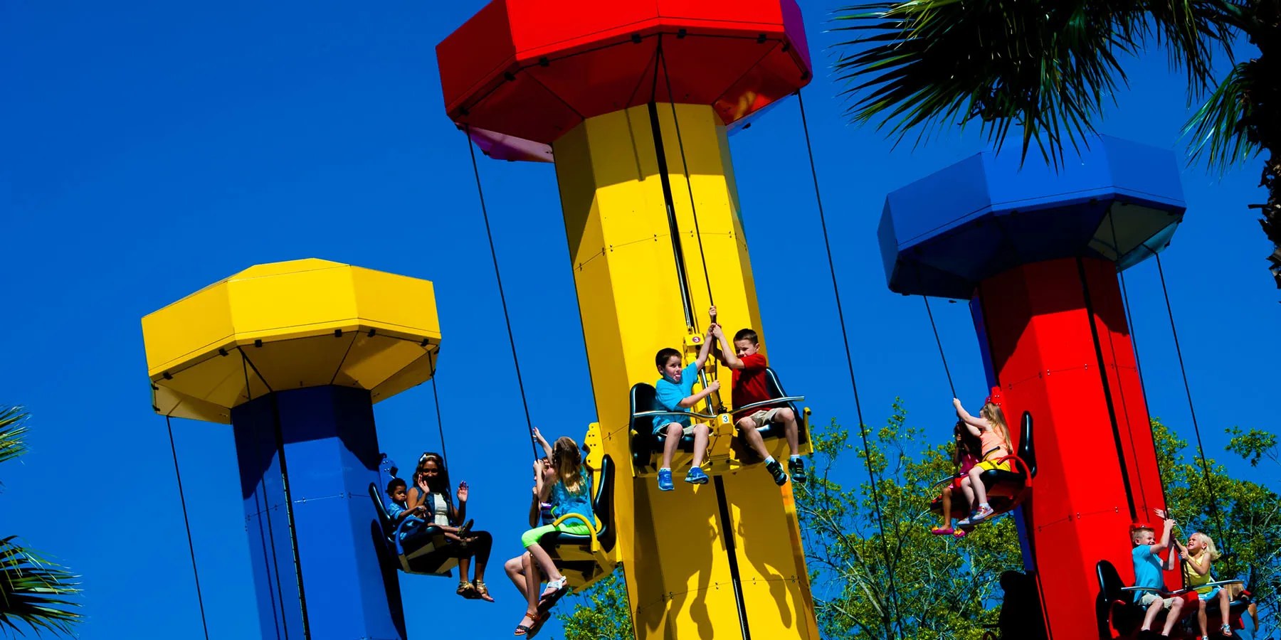 Children, and one adult riding with a small child, seated in pull-up ride seats hanging from three brightly colored LEGO brick styled towers against a deep blue sky with palm fronds