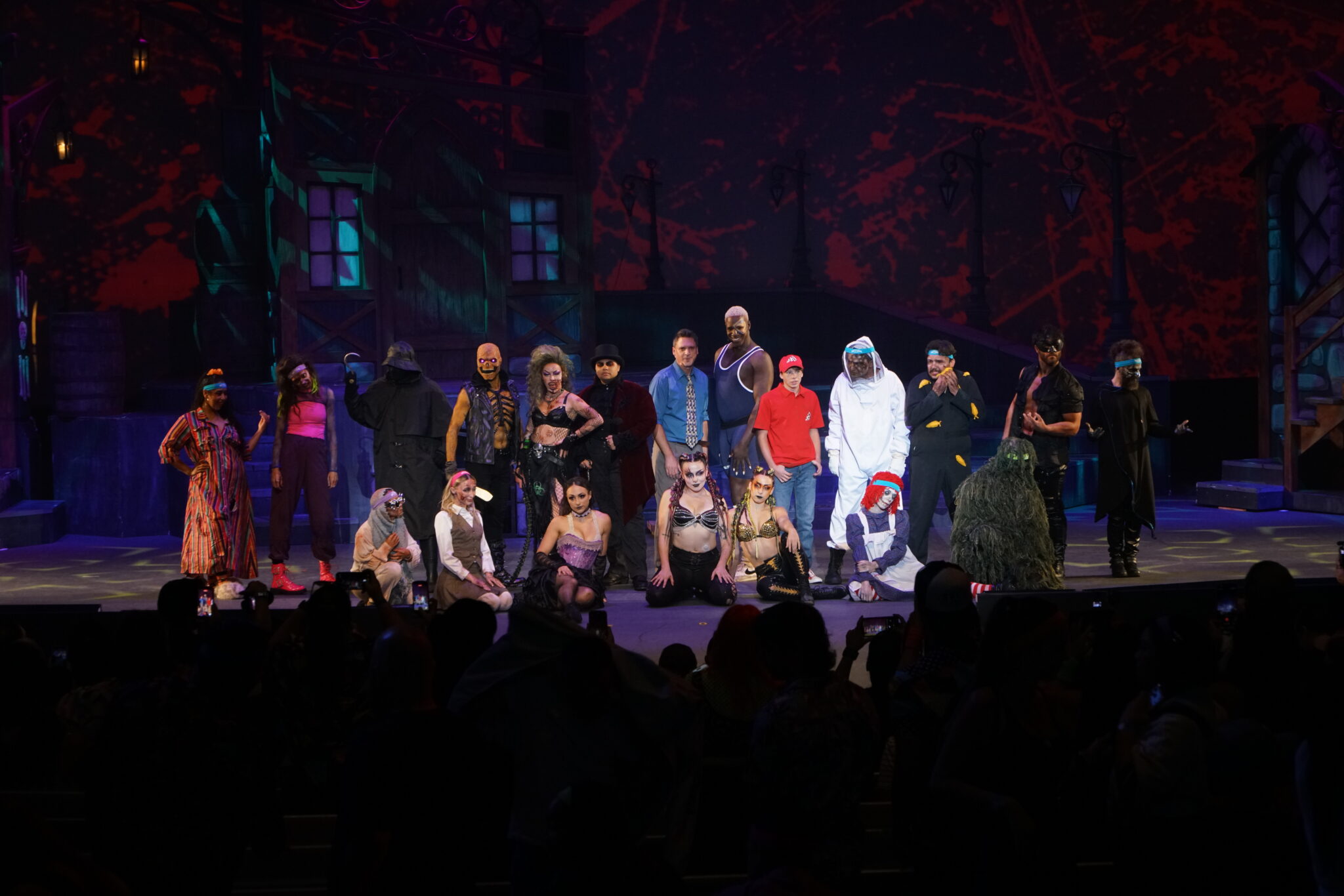 About twenty Howl-O-Scream performers in costume lined up across a theater stage in front of a red blood splatter backdrop, with audience silhouettes filming below