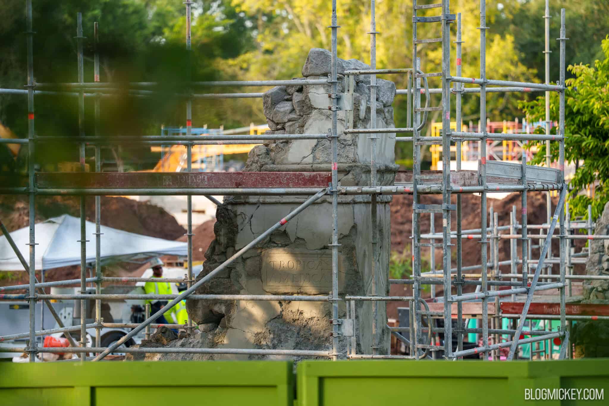 Wide view of a scaffolded stone column carrying the Tropical Americas tablet, seen over a lime green construction wall with a crew member and heavy equipment behind it