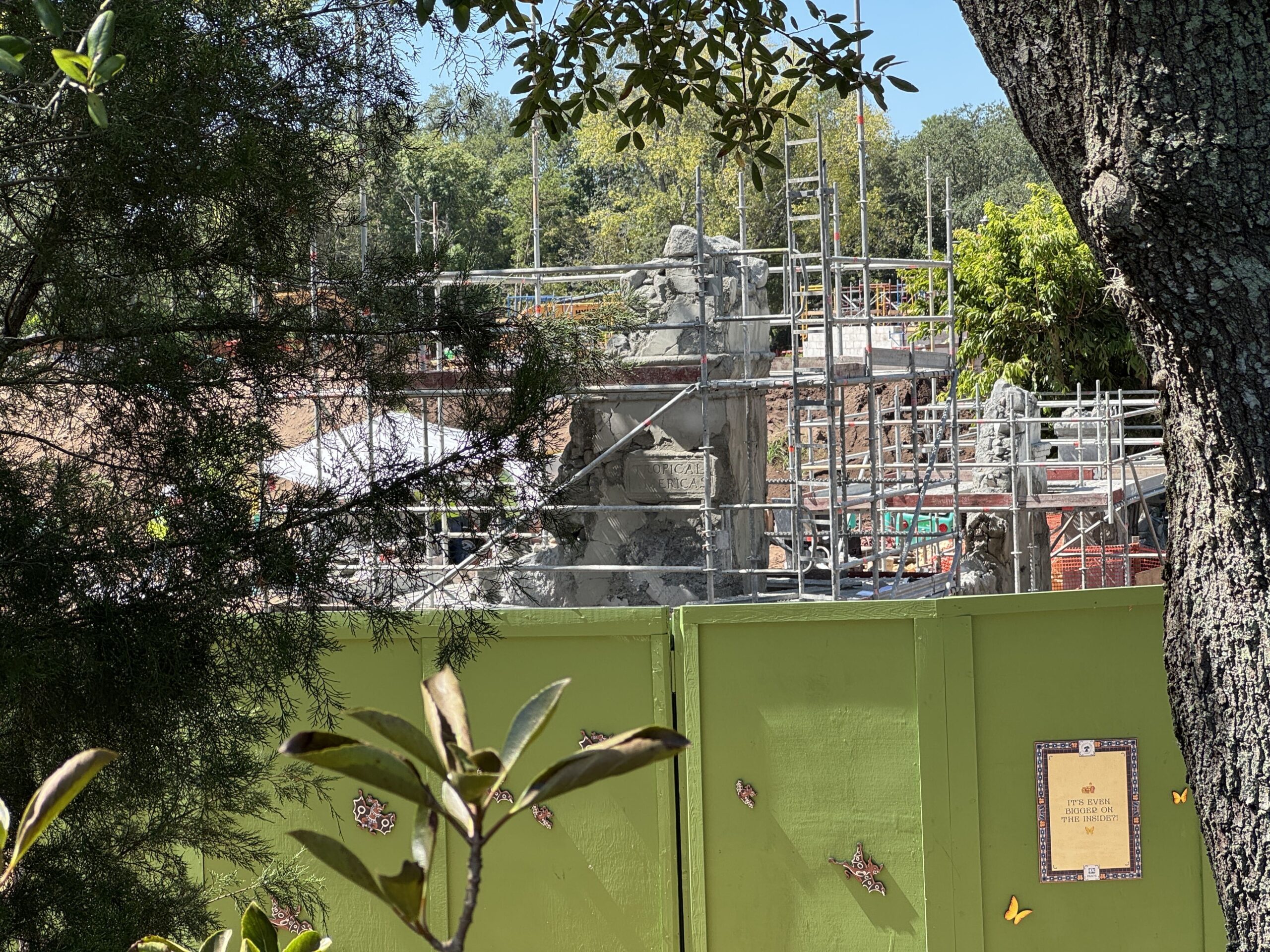 Guest-eye view from a park walkway framed by tree branches, looking over a lime green construction wall at the scaffolded secondary entrance column with the Tropical Americas tablet