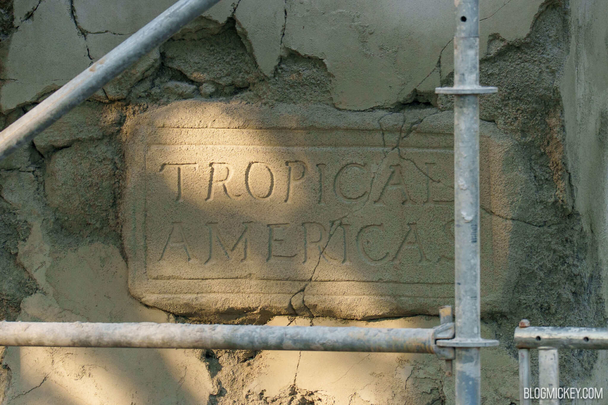 Close view of a carved stone tablet reading TROPICAL AMERICAS on a block standing proud of a column face, with cracked plaster around it and scaffolding poles crossing the frame