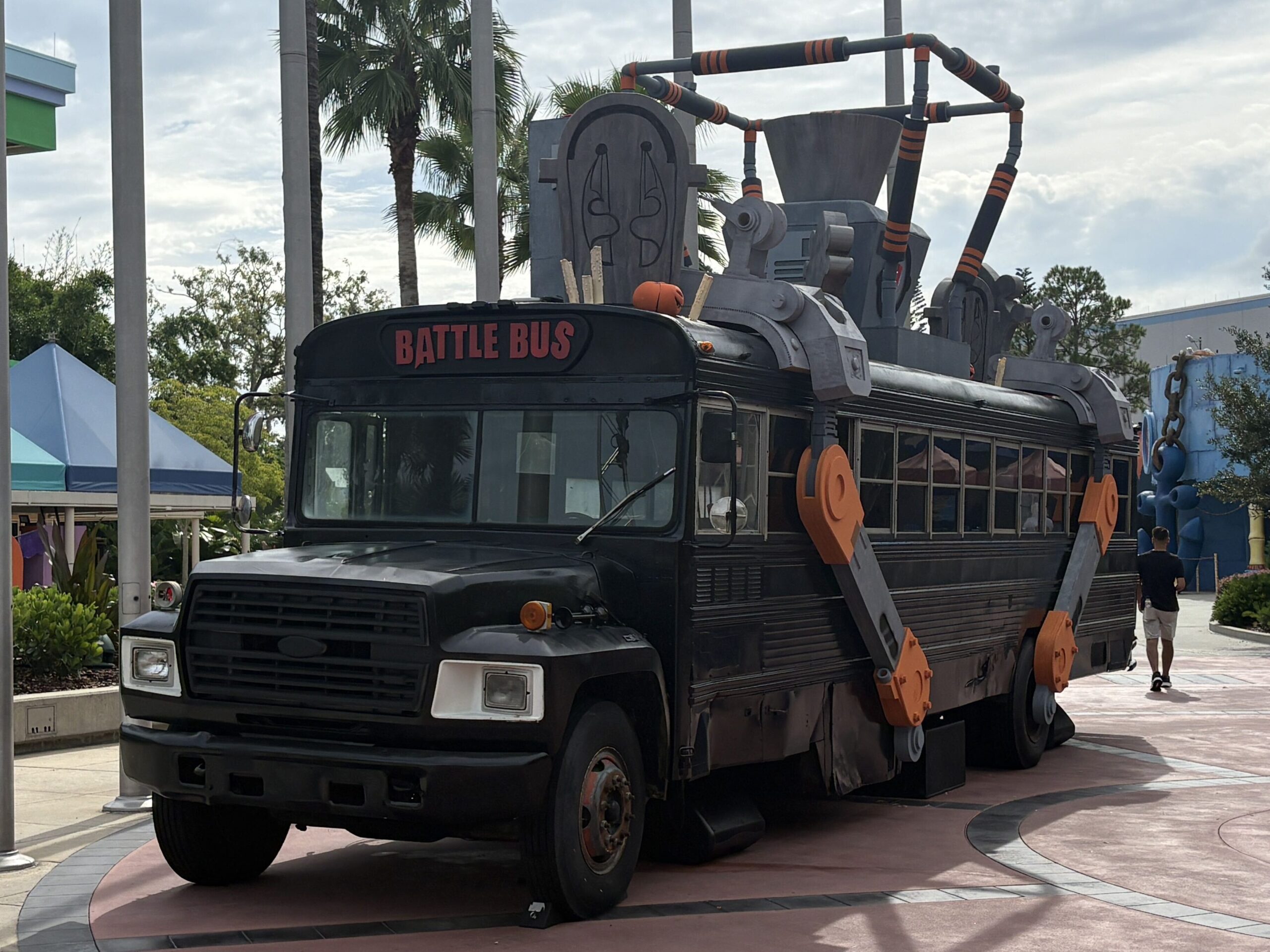 A matte black school bus lettered BATTLE BUS in red, with orange mechanical arms on its side and tombstones on its roof, parked on pink pavement between palm trees