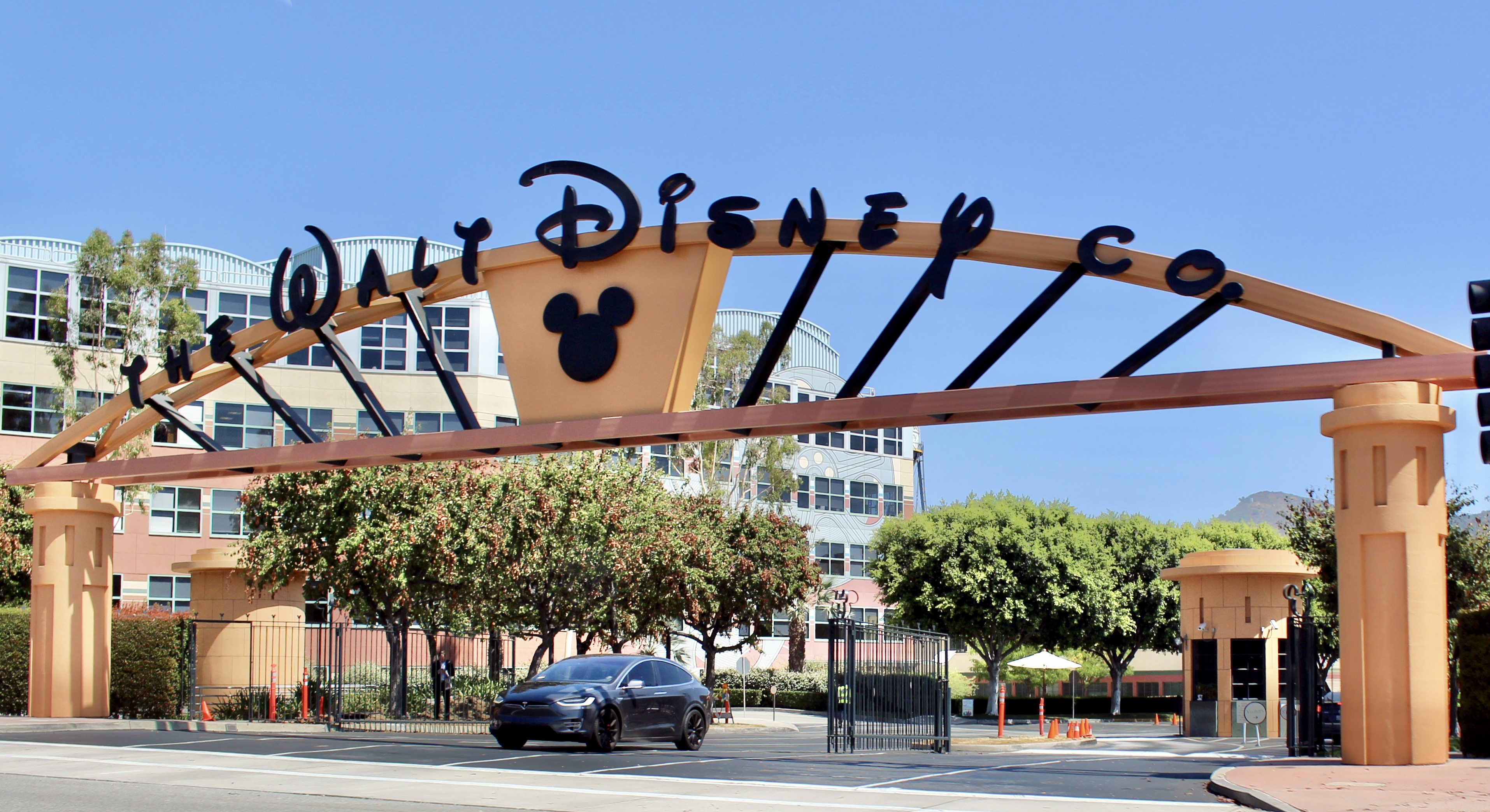 A curved orange beam spelling The Walt Disney Co. in black script letters over a road, with a black Mickey head on a gold panel at its center