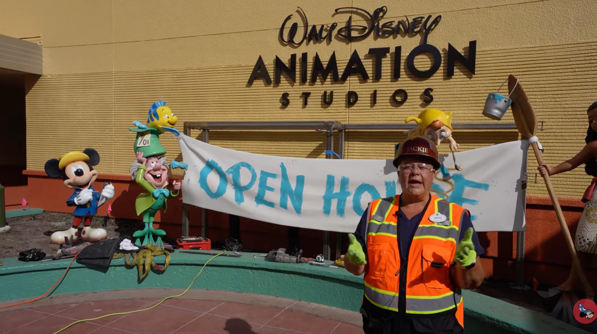 A Disney cast member in an orange safety vest and a hard hat reading Jackie stands in front of the Open House banner, with tools spread along a green planter wall and the statues in place