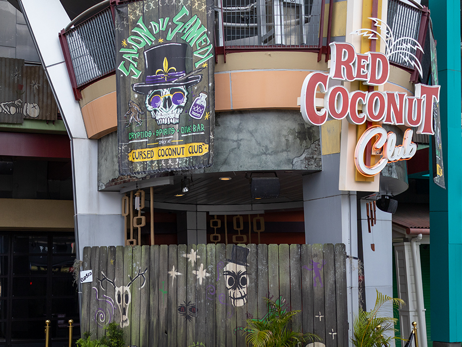 The exterior of the Red Coconut Club by day with a large painted Mardi Gras overlay banner hung beside the permanent neon sign, the banner reading SALON DU SAMEDI above a top hatted skull in purple sunglasses and CRYPTIDS SPIRITS DIVE BAR and ONLY AT CURSED COCONUT CLUB below it, with a weathered wooden fence painted with skulls in front