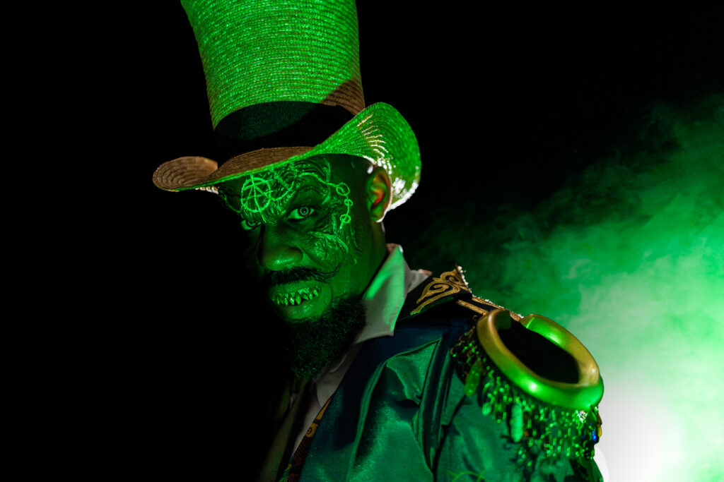 Dr. Oddfellow in close-up, lit entirely in green against black with green smoke drifting behind his shoulder, wearing a tall straw top hat, with glowing filigree markings across his face and an ornate gold shoulder piece hung with beaded fringe.