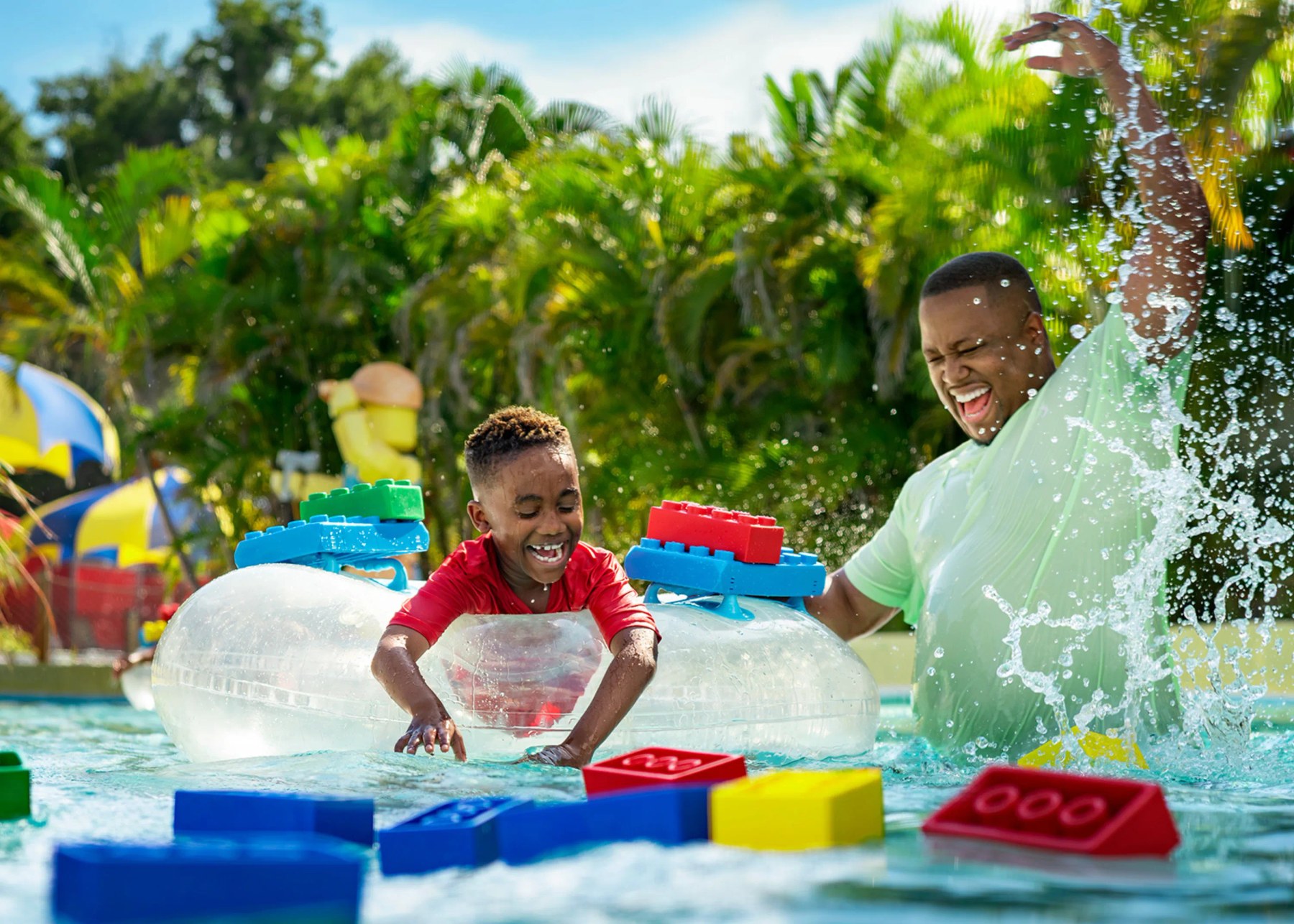 A boy in a red shirt in a clear tube and a man in a green shirt splashing beside him, with large soft LEGO bricks floating in the water