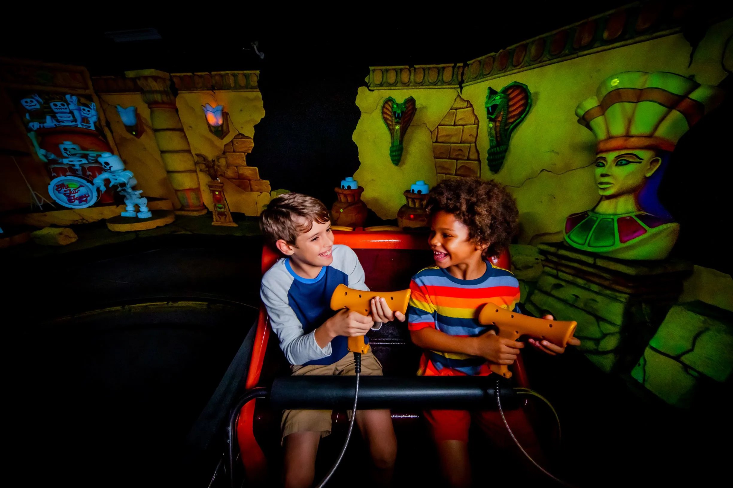 Two young boys sitting side by side in a red ride vehicle inside a dark show building on Lost Kingdom Adventure, each holding a chunky orange laser blaster on a coiled cable, turned toward each other laughing, with blacklit Egyptian tomb scenery behind them including a green lit pharaoh queen bust, two green cobras mounted on a cracked sandstone wall and a glowing LEGO skeleton panel