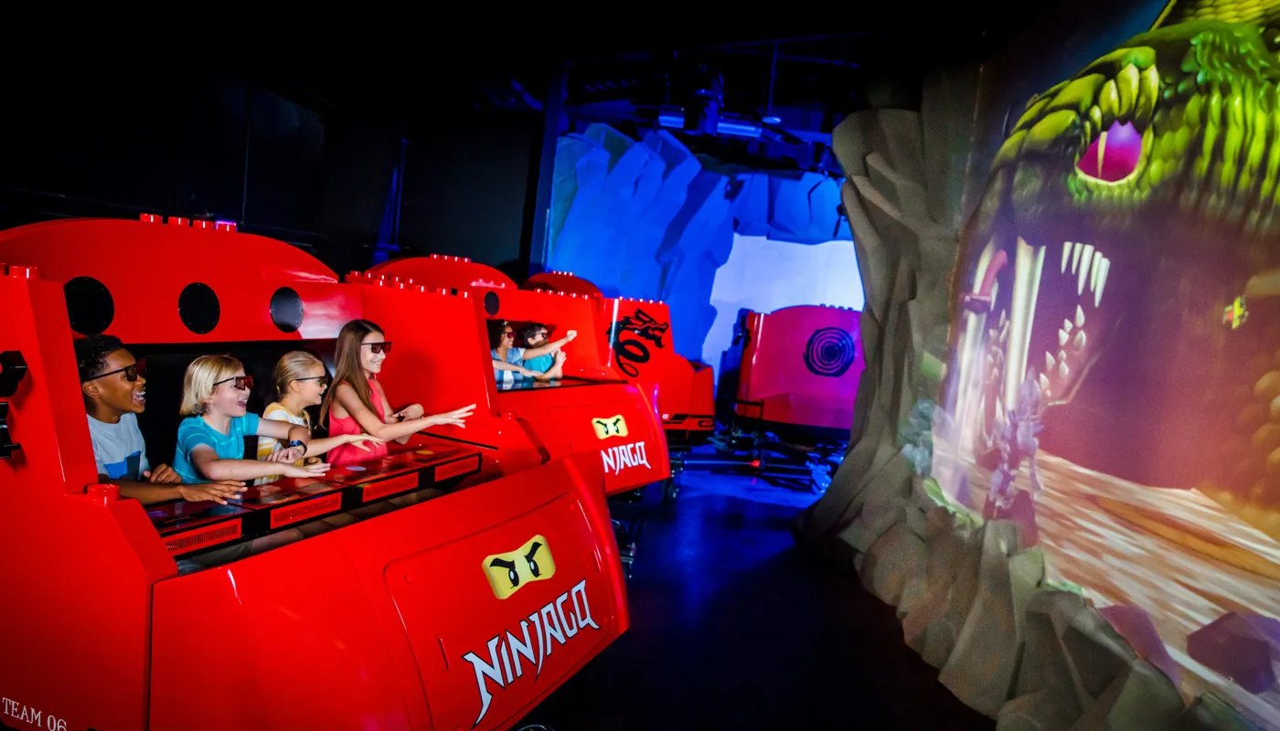 A train of red LEGO brick moulded ride cars on LEGO NINJAGO The Ride inside a dark show building, four children in the front car wearing 3D glasses and leaning forward with hands raised toward a large projected green serpent with an open mouth filling the right side of the frame, the word NINJAGO and a yellow ninja eyes decal printed on the side of the car