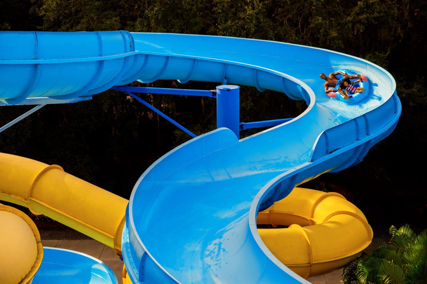 Two riders in a raft coming around a bend on a large blue water slide, with a yellow slide below and trees behind