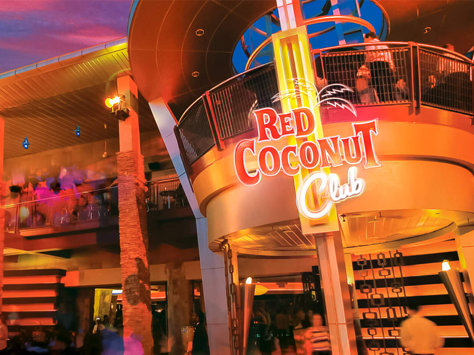 The Red Coconut Club at Universal CityWalk photographed from below at dusk, showing its curved second floor balcony with guests leaning on the railing, a large neon sign reading RED COCONUT CLUB in red and orange script, warmly lit stairs and columns, and a purple and pink sky behind