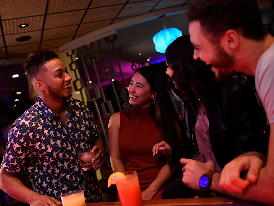Four guests laughing together around a bar height table inside the Red Coconut Club, two tall tropical cocktails in the foreground, one pale orange and one red, with teal and magenta lighting and a glowing blue pendant lamp behind them