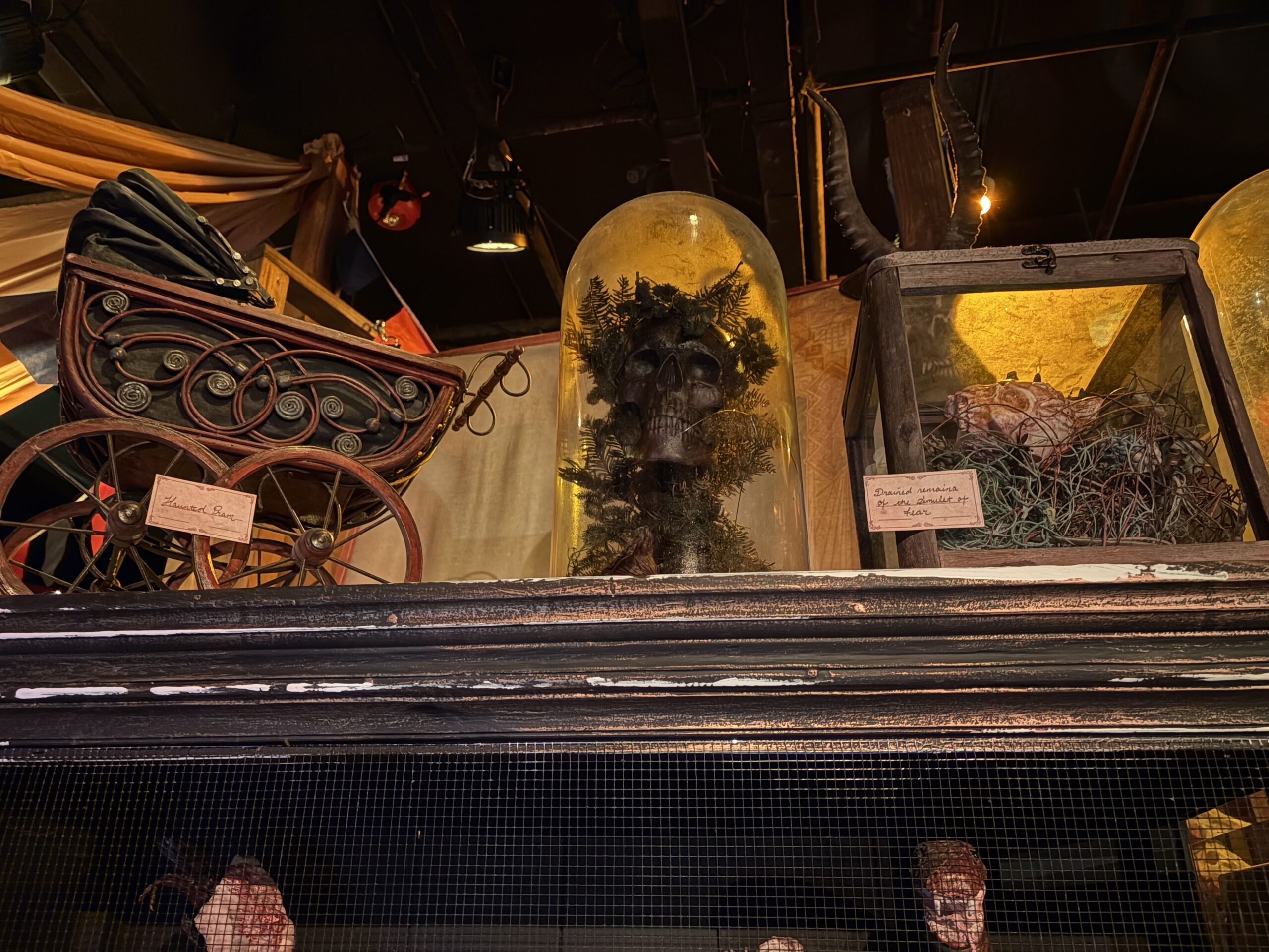 A Victorian pram labeled Haunted Pram beside a glass dome holding a skull in ferns, and a wooden case labeled Drained remains of the Amulet of Fear