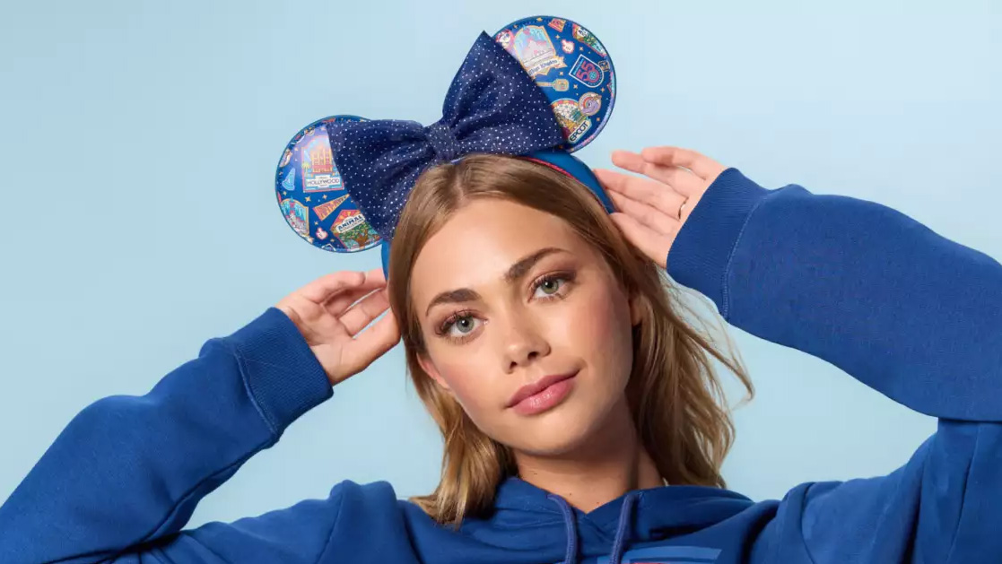 A model in the royal blue hoodie holding both hands up to a navy ear headband printed with colorful park icon badges and with a navy polka dotted bow at the center.