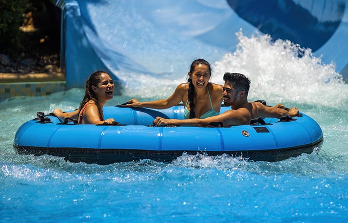 Three riders laughing in a large blue circular raft as it splashes out of the bottom of a white and blue water slide into a pool, water spraying up around the raft
