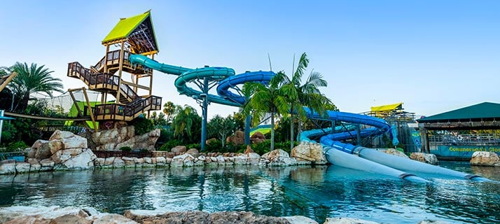 A water park slide complex under a bright pale blue daytime sky, with a wooden tower under a green and yellow roof at left, teal and blue flumes curving down beside it, a large white enclosed tube dropping from those flumes and running out low across a rock lined lagoon at center right, rockwork and palms around the water and a green roofed building at far right