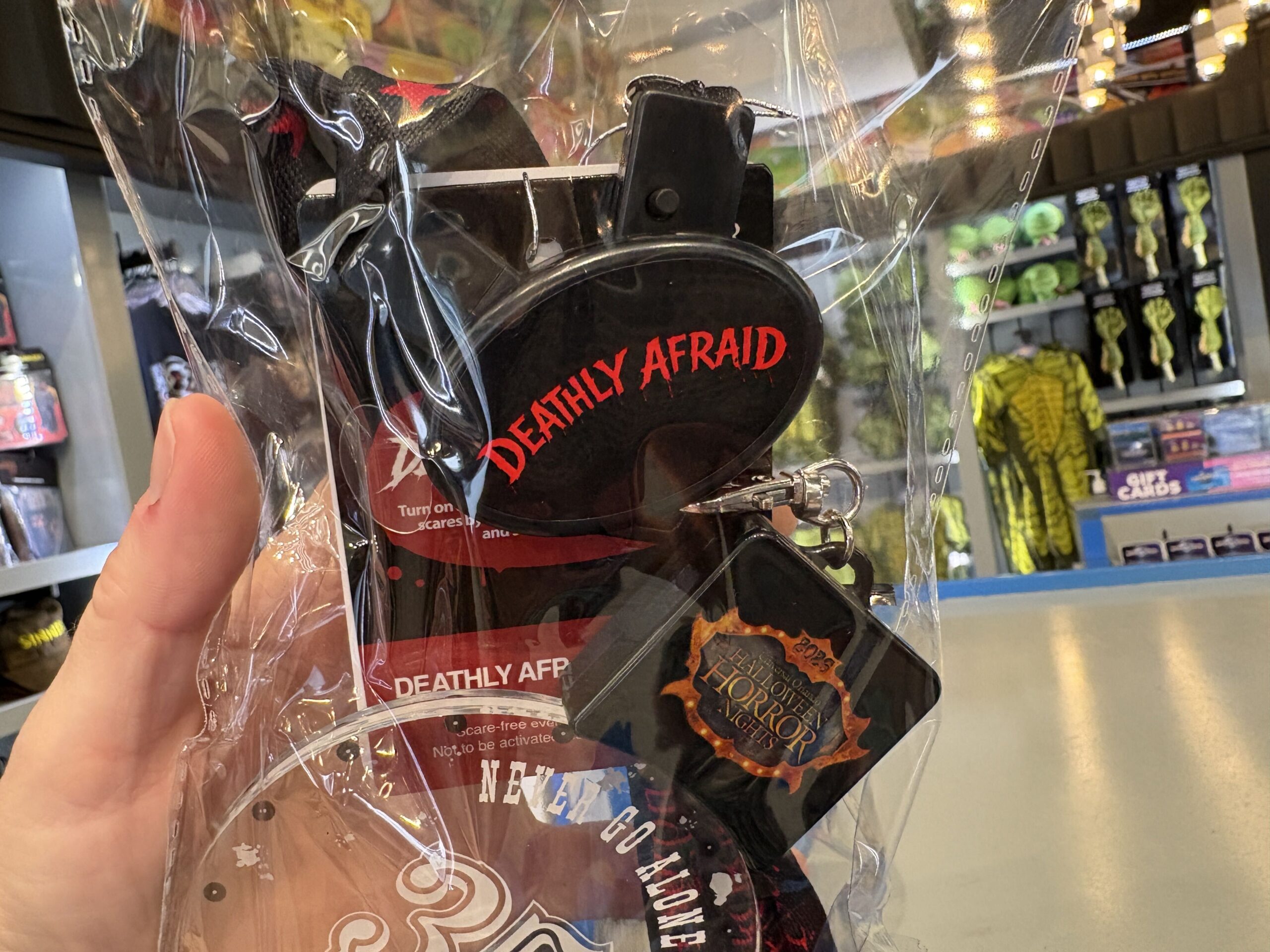 A packaged Deathly Afraid necklace held in a hand, showing a black oval piece with Deathly Afraid in dripping red letters and a black square tag bearing the Halloween Horror Nights crest
