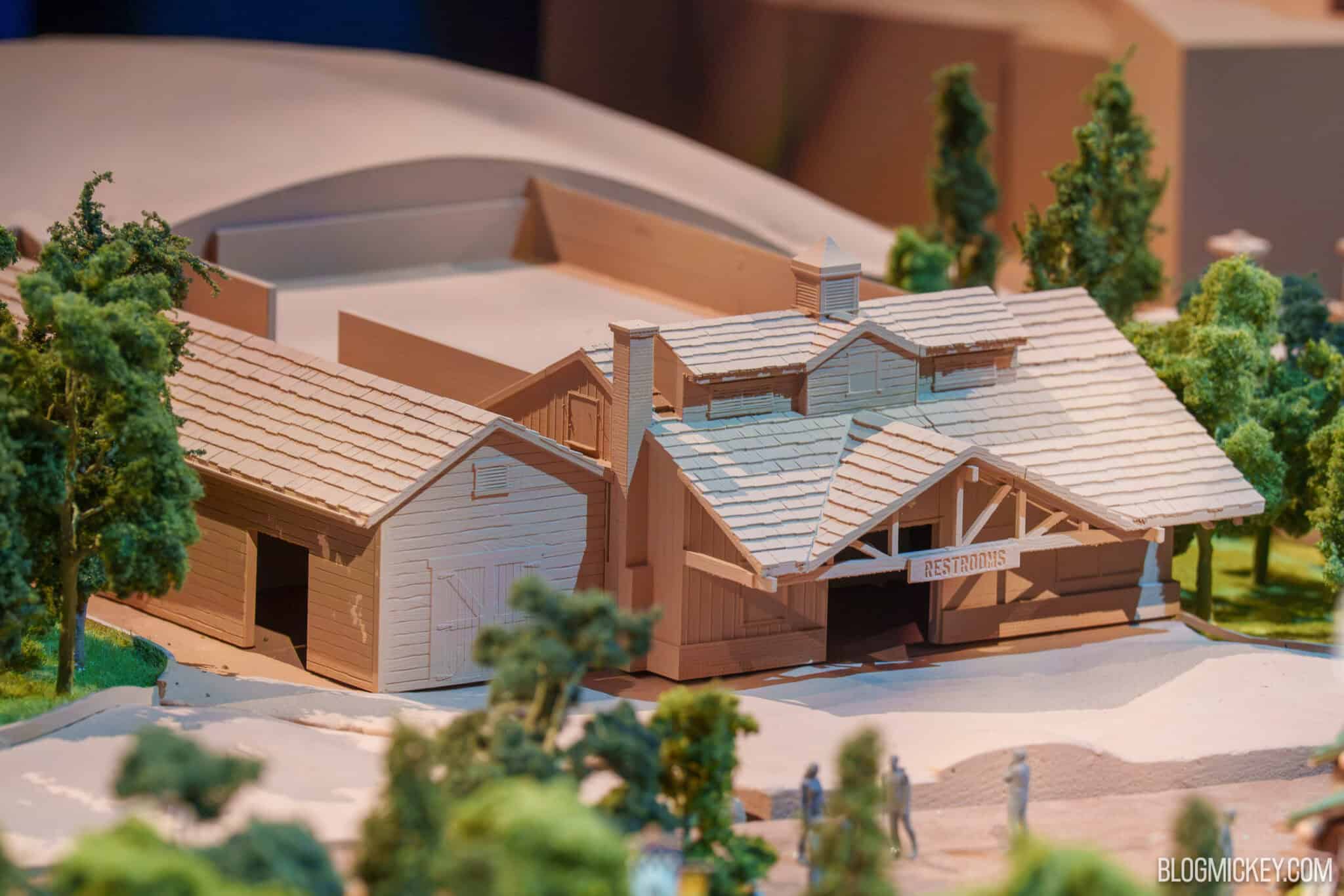A close view of an unpainted tan and cream architectural model building with a shingled roof, a cupola and an open timber porch, a small sign over the porch reading RESTROOMS, model trees on both sides and tiny gray figures walking past at lower right