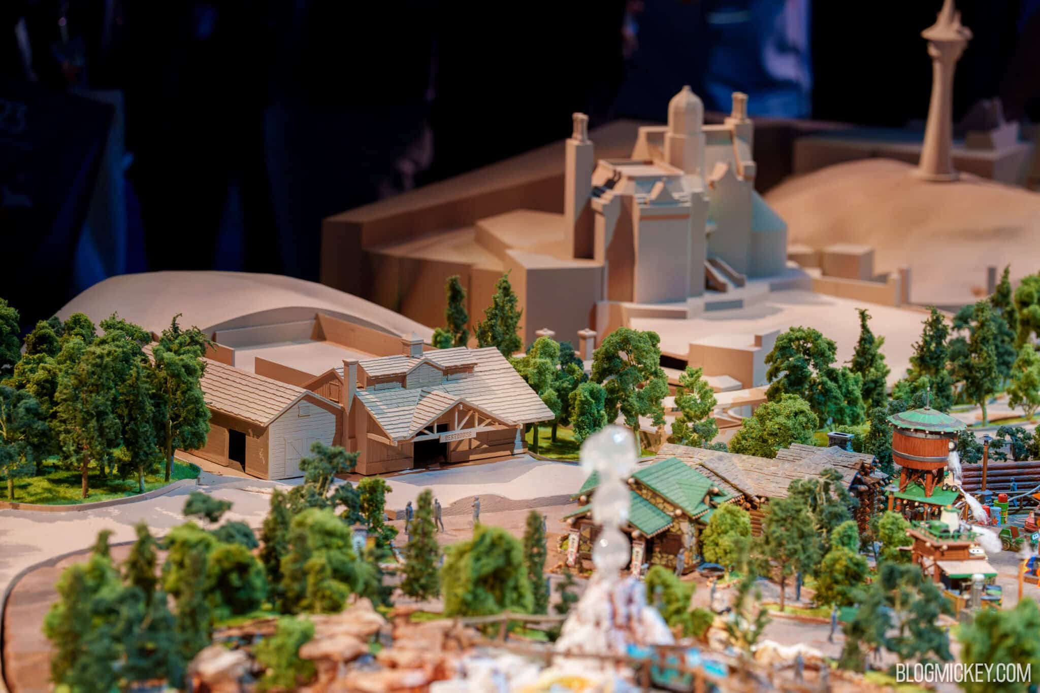 A wider view of the same architectural model showing the tan restroom building at left among model trees, a large unpainted pale massing with tall chimney stacks behind it, and fully painted green roofed log cabins, a wooden water tower at right and a white geyser plume in the foreground, against a dark exhibition hall background