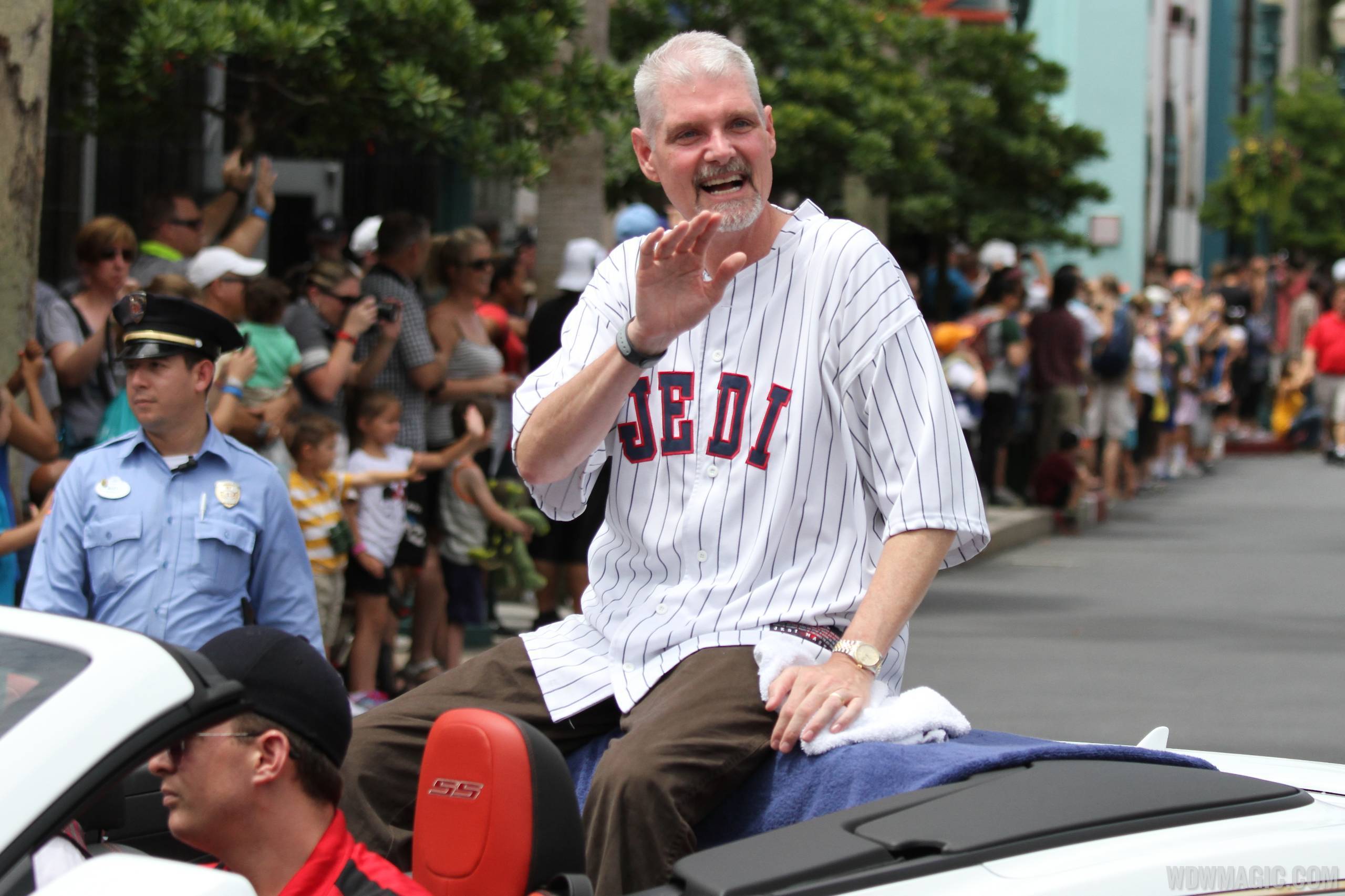 Tom Kane, a grey haired man with a goatee wearing a white pinstriped JEDI baseball jersey, waves from the back of a white convertible as a crowd lines the street behind him