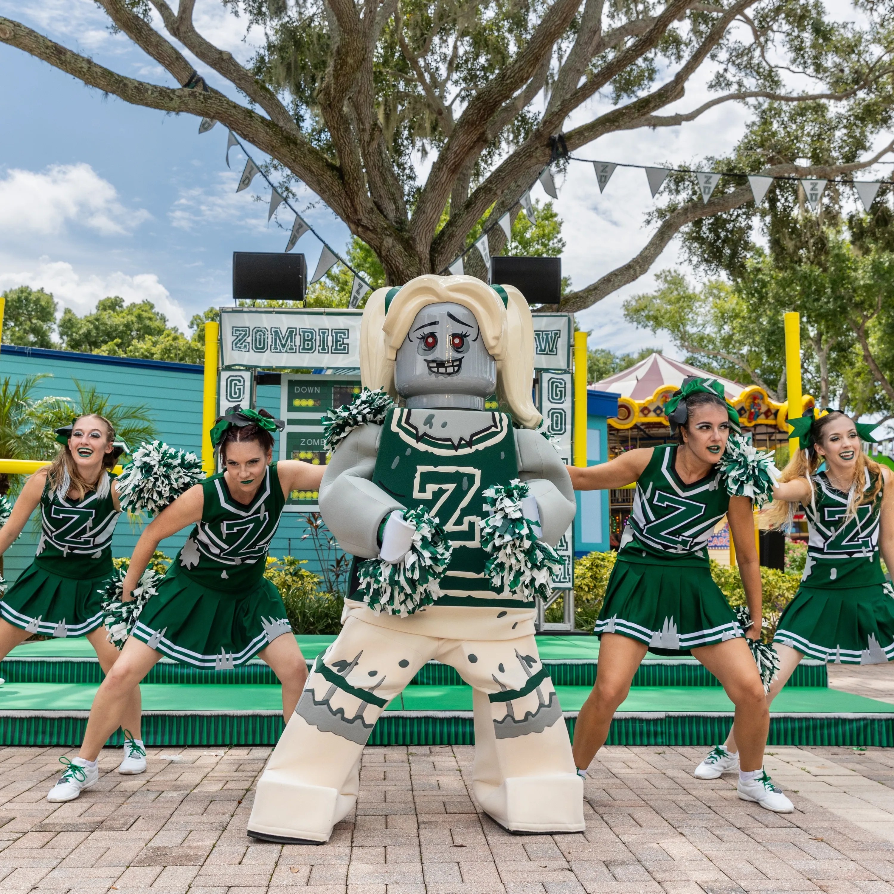Five human dancers, one partly cropped at the left edge, in dark green and white cheerleader uniforms with a large letter Z on the chest, holding green and white pom poms in a wide stance, flanking an oversized gray LEGO zombie cheerleader character in a matching green Z jersey and cream trousers, on brick pavement in front of a low green stage under a live oak with a ZOMBIE banner and scoreboard behind and a carousel at right