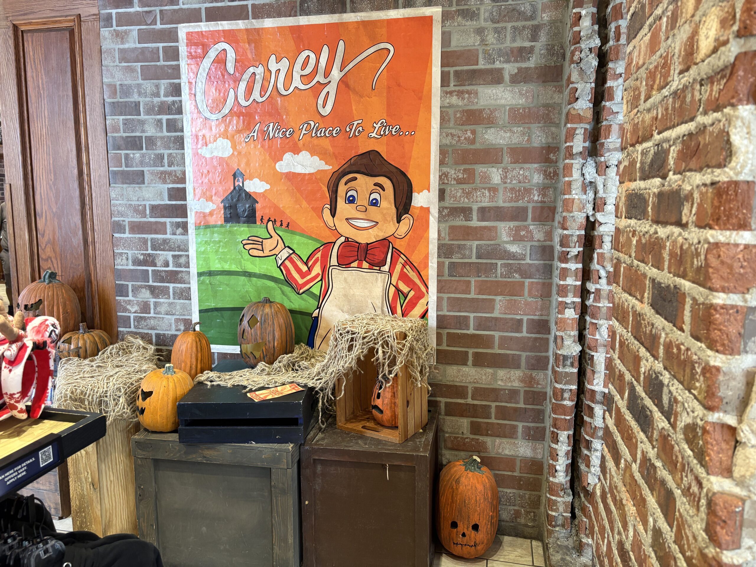 An orange Carey A Nice Place To Live poster on a brick wall showing a cartoon boy in an apron and bow tie gesturing toward a schoolhouse, with carved pumpkins below