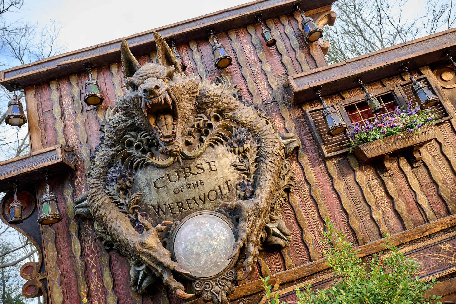 The carved wooden entrance marquee for Curse of the Werewolf, a snarling werewolf wrapped around a stone plaque holding a glowing orb, mounted on a red timber building hung with lanterns.