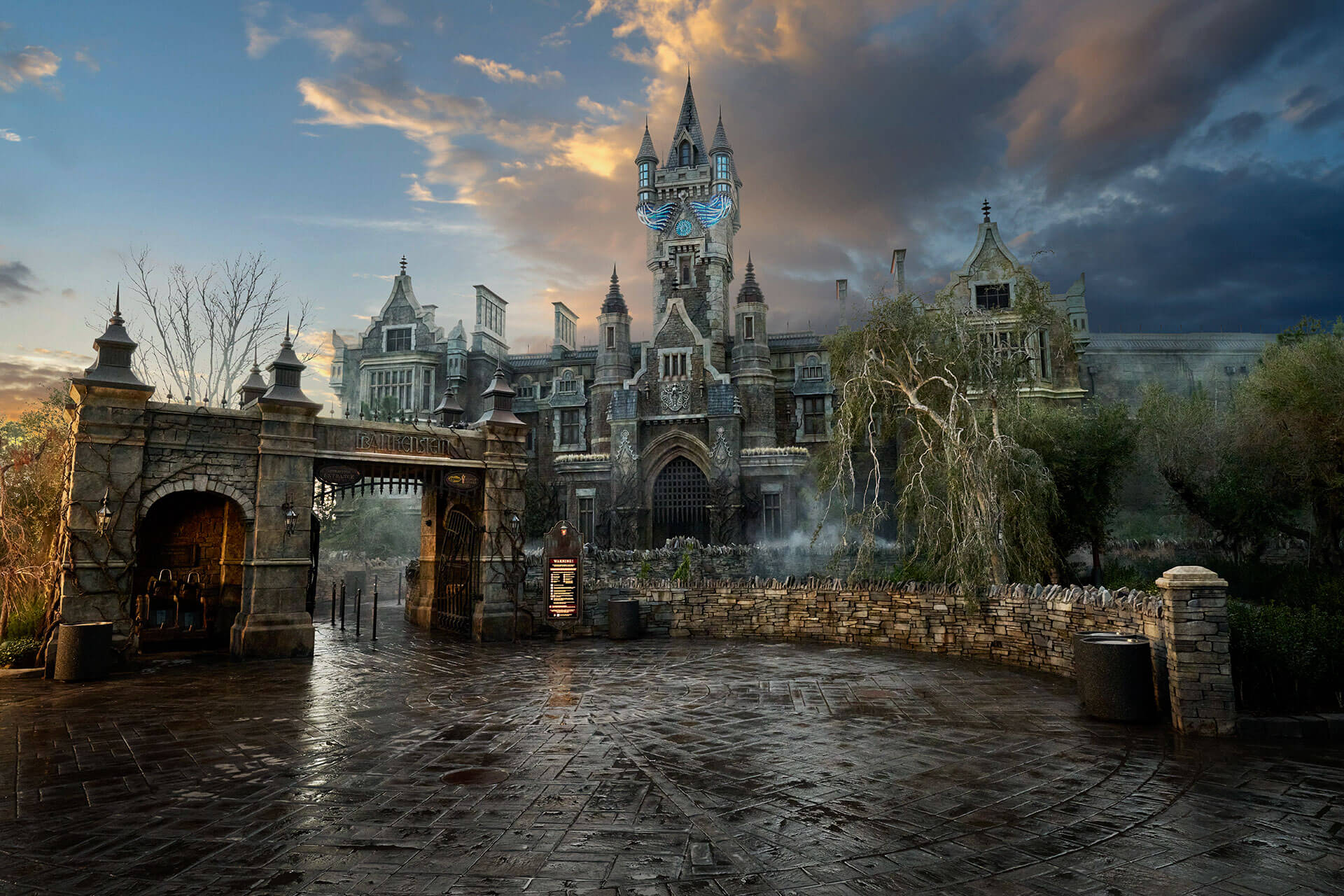 Frankenstein Manor at dusk, a tall gray Gothic mansion in Dark Universe at Epic Universe, with lit spires behind a stone gate arch reading Frankenstein, with wet paving in the foreground.