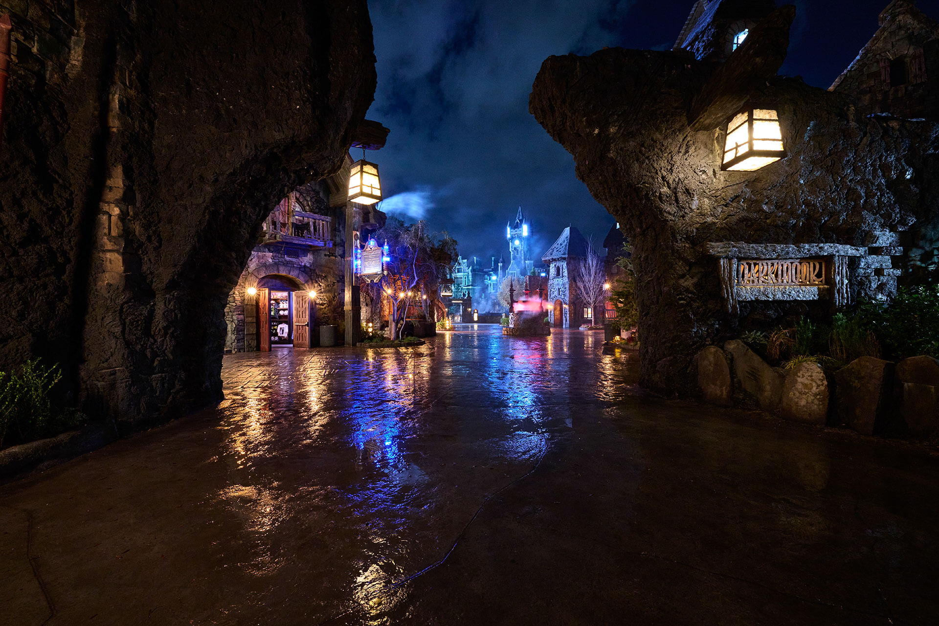 A wet cobblestone street in Darkmoor at night, framed by rocky outcrops, with glowing lanterns, a carved DARKMOOR sign at right and the illuminated Frankenstein Manor in the distance.