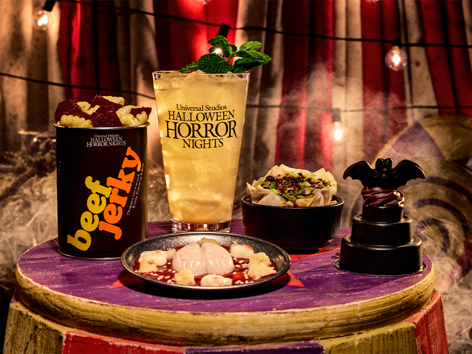 Five Halloween Horror Nights 35 menu items on a purple carnival barrel top: a black beef jerky can filled with pasta, a tall iced drink in an HHN cup garnished with mint, a black bowl of pot stickers topped with scallions and chili crisp, a plate holding a pale molded brain in red sauce with white cake pieces, and a small three tiered black cake with a chocolate bat on top