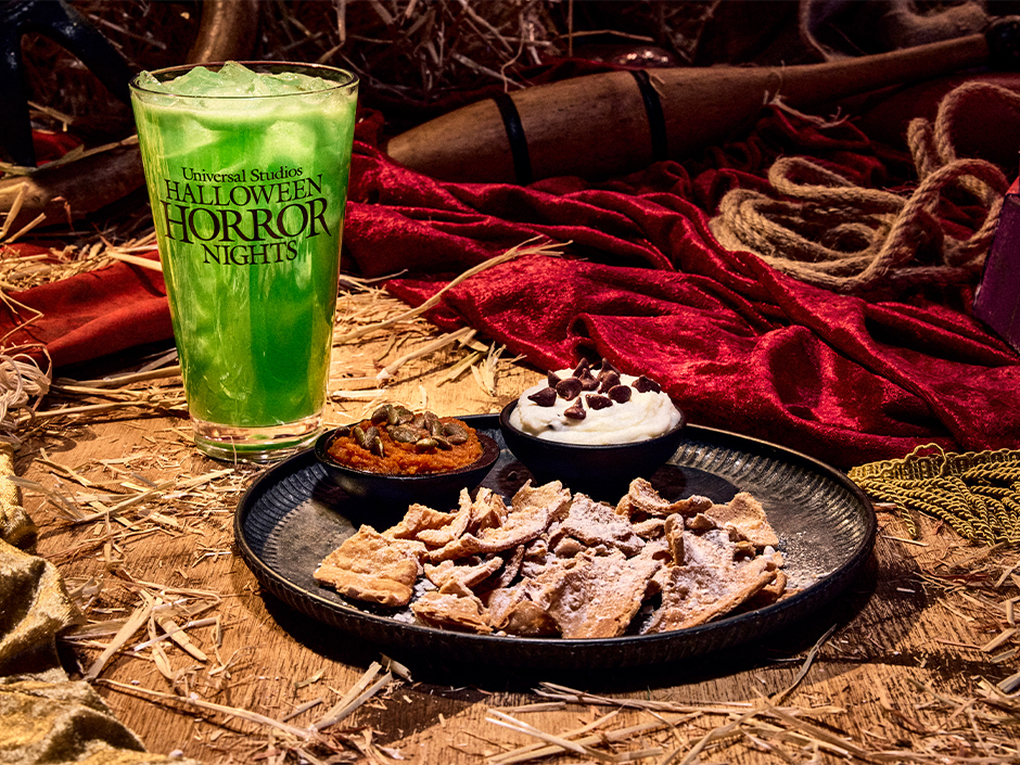 A bright green iced drink in a Halloween Horror Nights cup beside a black plate of powdered cannoli shell shards with two small bowls of dip, one orange pumpkin filling topped with pepitas and one white cannoli cream topped with chocolate chips, on a wooden surface scattered with straw and red fabric