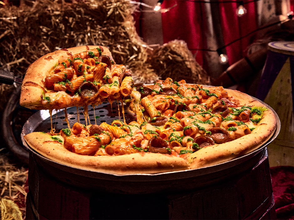 A whole pizza on a metal pan set on a wooden barrel, topped with baked rigatoni, sliced sausage and green pesto drizzle, with one slice being lifted away trailing melted cheese, in front of hay bales and a red carnival curtain