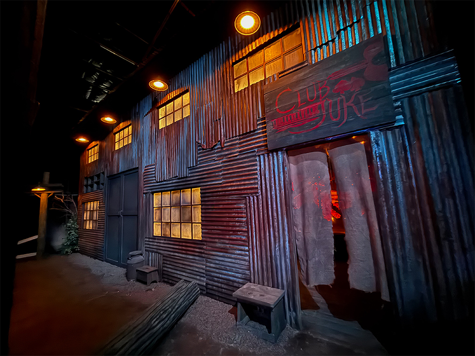 The Club Juke facade inside the Sinners haunted house at Halloween Horror Nights 35. A two story corrugated tin building with warm lit windows, a hanging red Club Juke sign, and a curtained doorway glowing red, with a bench and a fallen log in the dirt out front.