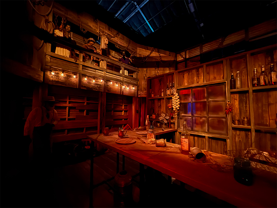 The interior of Club Juke inside the Sinners haunted house at Halloween Horror Nights 35. A warm lit wooden room with a long bar, bottles and jars on shelves, a string of bulb lights along a balcony rail, and a figure in a hat standing at the far left end of the bar.