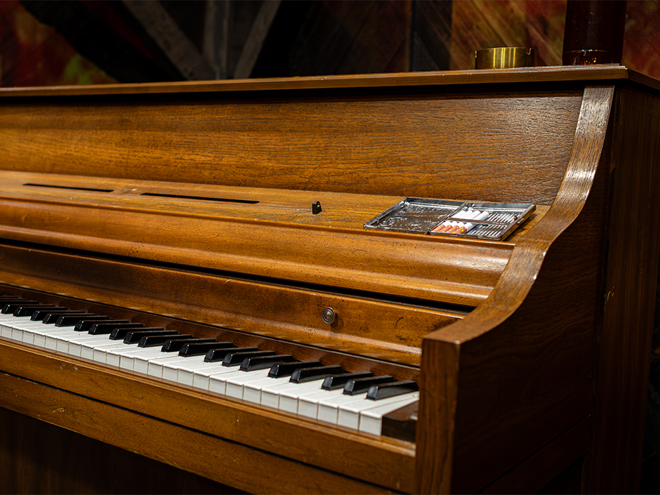 A close up of an upright wooden piano inside the Sinners haunted house at Halloween Horror Nights 35, with a metal ashtray holding cigarettes resting on the closed key cover ledge.